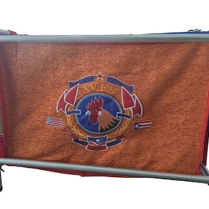 May include: A red, white, and blue embroidered logo with a rooster and the text "AVES" on an orange fabric backdrop. The fabric is stretched over a metal frame.
