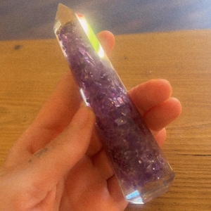 May include: A clear, hexagonal, wand-shaped object made of resin, filled with small, deep purple amethyst-colored stone fragments. The object has a pointed, faceted top and a smooth, cylindrical body that reflects light along its edges.