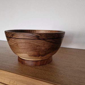 Könnte beinhalten: Handgefertigte Holzschale in dunkler Braunfärbung. Die Schale hat eine runde Form und steht auf einem kreisförmigen Sockel. Die Holzmaserung ist sichtbar, was zur natürlichen Ästhetik beiträgt. Durchmesser ca. 25 cm. Ideal zum Servieren oder Dekorieren.
