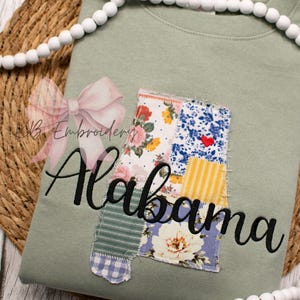May include: Sage green sweatshirt with the embroidered word "Alabama" in black script. The state outline is patched with floral and patterned fabrics. A pink bow and white beaded garland are in the background.