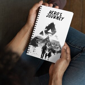May include: A white spiral notebook with a black and white illustration of a family walking through a forest. The notebook has the text "HERO'S JOURNEY" printed on the cover.