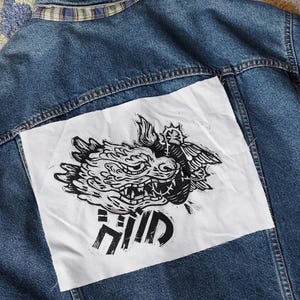 以下が含まれることがあります： 後ろに白いパッチが縫い付けられた青いデニムジャケット。パッチには、翼のある生き物の白黒のイラストと、その下に「MIND」という文字が印刷されています。