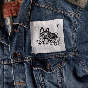 以下が含まれることがあります： 白のパッチが付いた青いデニムジャケット。パッチには、スタイリッシュな動物の頭部の白黒のイラストが描かれています。