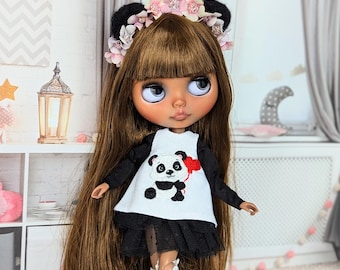 May include: A doll with long brown hair, wearing a white dress with a panda design and a black skirt. The doll has a floral headband with black bear ears and white knee-high boots. The doll's eyes are large and blue.