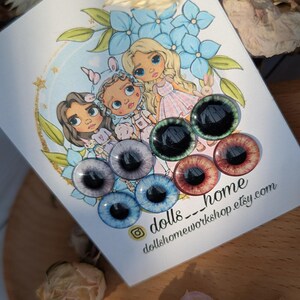 May include: A set of six colorful doll eyes with black pupils. The eyes are arranged in a circle on a white card with a drawing of three dolls and the text "dolls_home dollshomeworkshop.etsy.com".