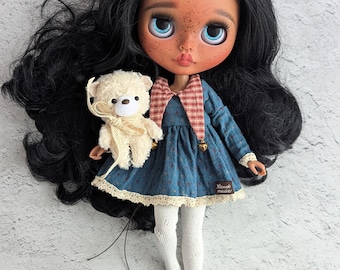 May include: A handcrafted doll with black hair and blue eyes, wearing a blue dress with a red and white plaid collar and white tights. The doll holds a small, cream-colored teddy bear. The doll's face has freckles and a soft, painted expression.