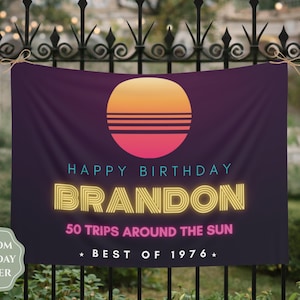 Op de afbeelding: Een rechthoekige donkerpaarse verjaardagsbanner met een zonsondergangafbeelding met oranje en roze horizontale strepen. De tekst luidt HAPPY BIRTHDAY BRANDON in lichtblauw en geel, gevolgd door 50 TRIPS AROUND THE SUN en BEST OF 1976.