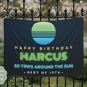 Op de afbeelding: Een donkerblauwe rechthoekige verjaardagsbanner met een cirkelvormige afbeelding in een groen naar blauw verloop en horizontale lijnen. De tekst luidt: HAPPY BIRTHDAY MARCUS 50 TRIPS AROUND THE SUN BEST OF 1976. De banner is aan de bovenhoeken met touw bevestigd.
