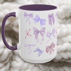 May include: A white ceramic mug with a purple rim and handle. The mug features a pattern of watercolor bows in shades of purple, pink, and lavender.