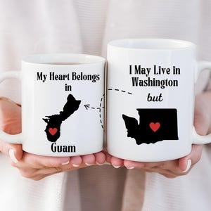 May include: Two white ceramic mugs with black silhouettes of Guam and Washington state, each with a red heart. One mug reads "My Heart Belongs in Guam," the other says "I May Live in Washington but."