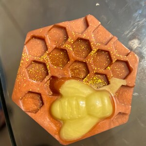 Puede incluir: Un jabón hexagonal con un diseño de panal y un acento amarillo en forma de abeja. El jabón es de color naranja cálido con detalles de purpurina dorada, creando un efecto brillante. Este jabón hecho a mano es perfecto para un regalo de spa.