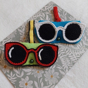 May include: Two crocheted eyeglass cases, one with a green and red design and the other with a blue and white design. Both cases have black crocheted lenses.