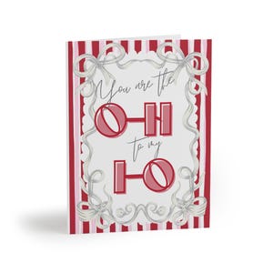 May include: A greeting card featuring a white center panel with red and pink vertical stripes on the sides. The card displays the text "You are the HO to my HO" in red, surrounded by a decorative gray ribbon border with bows at each corner.