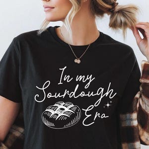 Op de afbeelding: Zwart T-shirt met de tekst "In my Sourdough Era" en een witte illustratie van een brood. Het shirt heeft korte mouwen en een ronde hals. De persoon draagt een bruin en tan geruit overhemd.