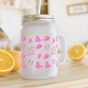 May include: White mason jar with a pink and white pattern of cowboy boots, hats, horseshoes, and the words "Yee Haw". The jar has a lid and a handle. It is sitting on a wooden surface with two oranges in the background.