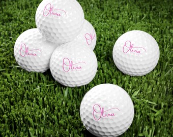 May include: White golf balls with the name "Olivia" printed in pink script. The golf balls are arranged on a green grass surface. The golf balls have a dimpled surface.