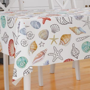 Può includere: Tovaglia bianca con un motivo a conchiglie e stelle marine. La tovaglia presenta illustrazioni ad acquerello di conchiglie, stelle marine e ciottoli in tonalità di blu, marrone, rosso e giallo.