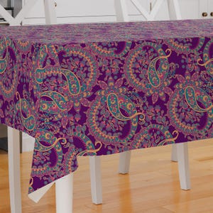May include: A rectangular tablecloth with a vibrant paisley pattern. The design features large, stylized floral motifs in shades of purple, teal, gold, and pink. The tablecloth is draped over a white table, showcasing the intricate details of the fabric.