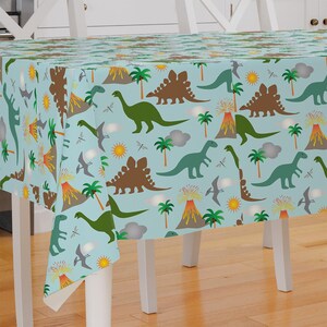 May include: A blue plastic tablecloth with a dinosaur pattern. The tablecloth features green and brown dinosaurs, palm trees, volcanoes, and clouds on a light blue background.