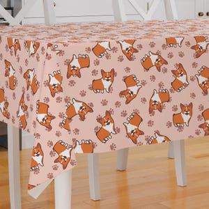 Peut inclure: Une nappe rose avec un motif répété d'empreintes de pattes brunes et de corgis de dessin animé. Les corgis sont orange et blancs avec des nez noirs et des visages souriants.