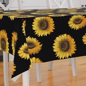 May include: Black tablecloth with a repeating pattern of yellow sunflowers with brown centres. The sunflowers are realistically depicted with detailed petals. The tablecloth is draped over a white table, contrasting the dark fabric with the light furniture.