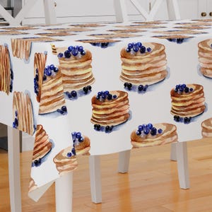May include: White tablecloth with a repeating pattern of watercolour pancakes topped with blueberries. The pancakes are stacked and have a light brown colour.