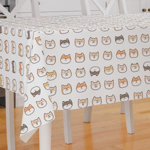 Peut inclure: Nappe blanche avec un motif répété de visages de chiens Shiba Inu bruns et orange. La nappe est drapé sur une table blanche avec quatre pieds blancs.