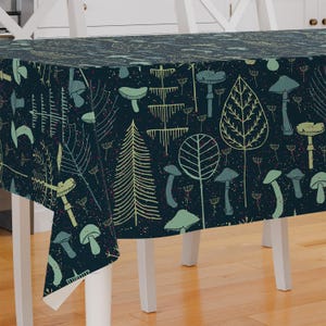 Peut inclure: Une nappe bleu foncé avec un motif répété de champignons, de feuilles et d'arbres stylisés dans des tons de vert et de beige. Le motif est imprimé sur le tissu, créant une esthétique fantaisiste et inspirée de la nature. La nappe est rectangulaire et recouvre une table blanche.