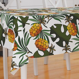 May include: White tablecloth with a tropical print featuring pineapples and green leaves. The tablecloth is laid on a wooden table.