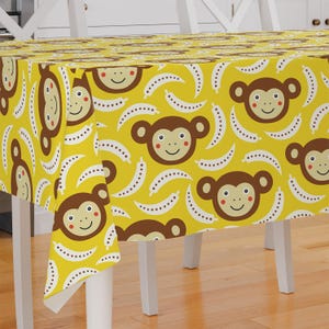Könnte beinhalten: Gelber Tischdecke mit einem sich wiederholenden Muster aus braunen Affen und weißen Bananenschalen. Die Affen haben braunes Fell, weiße Augen und rote Wangen. Die Bananenschalen sind mit weißen Punkten besetzt.