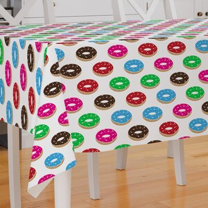 May include: White tablecloth with a repeating pattern of colorful donuts. The donuts are in shades of pink, red, green, blue, and brown. Each donut has white sprinkles.