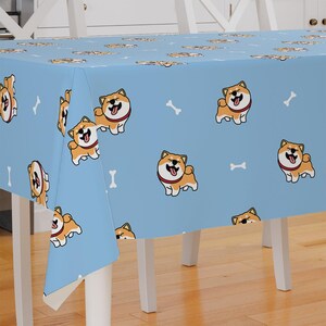 Peut inclure: Une nappe bleu clair avec un motif répété de chiens Shiba Inu dessinés et d'os blancs. La nappe est sur une table en bois avec des chaises blanches.