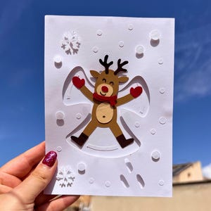 May include: A handmade Christmas card featuring a reindeer snow angel design. The reindeer has brown fur, black antlers, a red scarf and mittens. The card is white with embossed snowflakes and snow angel details.