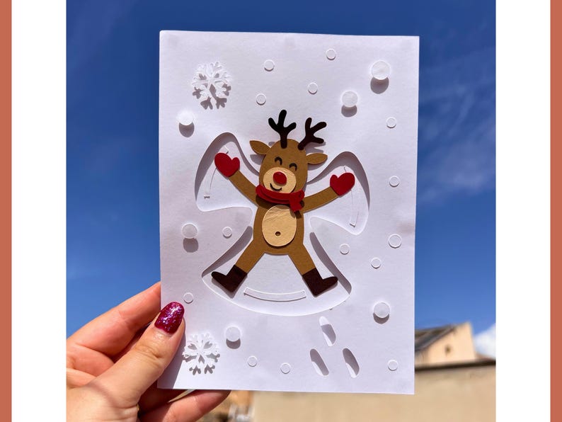 May include: A handmade Christmas card featuring a reindeer snow angel design. The reindeer has brown fur, black antlers, a red scarf and mittens. The card is white with embossed snowflakes and snow angel details.