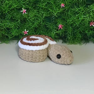 Puede incluir: Un juguete de peluche de crochet marrón y blanco con forma de rollo de canela. El juguete tiene ojos negros y un remolino marrón en la parte superior.