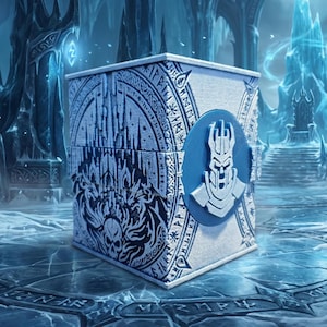 Op de afbeelding: Een vierkante witte doos met gedetailleerde zwarte lijntekeningen van een kasteel en een schedel, omringd door een cirkelvormige rand met runensymbolen. Een blauw rond embleem met een zilverkleurig harnashelm-icoon is in het midden geplaatst.