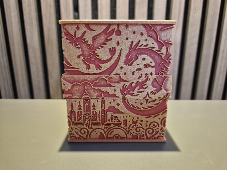 Peut inclure: Une bo&icirc;te rectangulaire rose avec des motifs d&eacute;coup&eacute;s au laser. La bo&icirc;te pr&eacute;sente des dragons, des &eacute;toiles et un paysage urbain. Le design est d&eacute;taill&eacute; et la bo&icirc;te semble &ecirc;tre en bois ou dans un mat&eacute;riau similaire. Le th&egrave;me g&eacute;n&eacute;ral est la fantaisie.