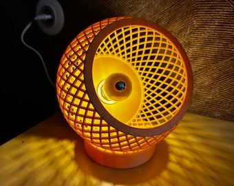 Könnte beinhalten: Eine moderne Tischlampe in Orange mit einem geometrischen, offenen Webmuster. Die Lampe hat einen runden Sockel und eine Glühbirne im Inneren.