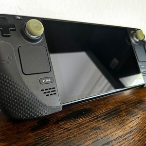 Może przedstawiać: Czarne przenośne urządzenie do gier z ekranem dotykowym, dwoma analogowymi gałkami i przyciskiem z napisem "STEAM".