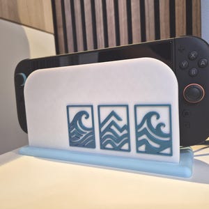 Può includere: Un supporto per console di gioco bianco e azzurro con tre disegni di onde blu. Una console di gioco nera è posizionata nel supporto. Il supporto è in plastica e poggia su una superficie bianca.
