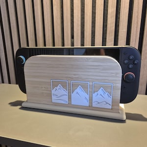 May include: A light-colored wooden game console stand with three mountain designs. A black and gray game console is placed in the stand. The stand is made of light-colored wood and has a rectangular base.