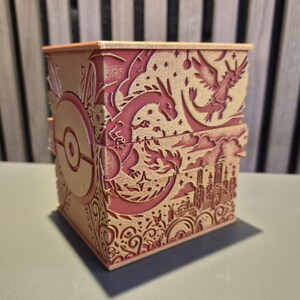 Peut inclure: Une bo&icirc;te d&eacute;corative bicolore rose et beige avec des motifs d&eacute;coup&eacute;s au laser complexes. La bo&icirc;te pr&eacute;sente un th&egrave;me Pok&eacute;mon, avec des images de dragons, d'une ville et d'une Pok&eacute; Ball. La bo&icirc;te est rectangulaire avec un couvercle.