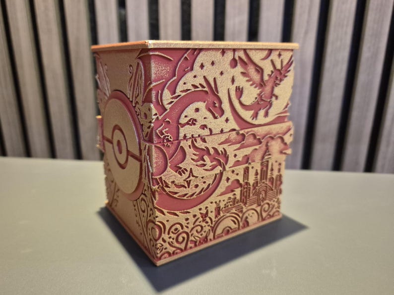 Peut inclure: Une bo&icirc;te d&eacute;corative bicolore rose et beige avec des motifs d&eacute;coup&eacute;s au laser complexes. La bo&icirc;te pr&eacute;sente un th&egrave;me Pok&eacute;mon, avec des images de dragons, d'une ville et d'une Pok&eacute; Ball. La bo&icirc;te est rectangulaire avec un couvercle.