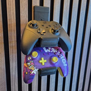 Op de afbeelding: Een zwarte en paarse videogamecontroller met een cartoonkarakterontwerp, gemonteerd op een houten muur. De controller heeft een kruisvormig richtingspad en gele knoppen. Het woord "Waluigi" is zichtbaar.
