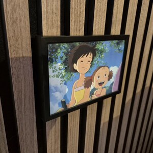 以下が含まれることがあります： 日本の映画に登場する2人のアニメキャラクターが描かれたフレームプリント。キャラクターは若い女の子と男の子で、青い空と緑豊かな緑を背景にしています。フレームは黒で、プリントは木の壁に取り付けられています。