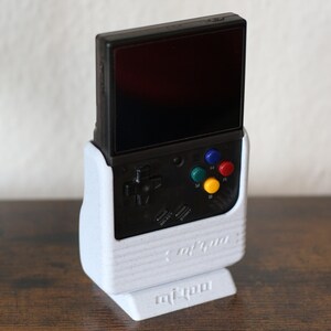 May include: A white stand with a black and gray handheld gaming console. The console has a screen, four colored buttons, and a directional pad. The stand has the text "miSTer" printed on the bottom.