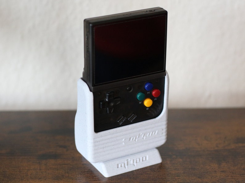 May include: A white stand with a black and gray handheld gaming console. The console has a screen, four colored buttons, and a directional pad. The stand has the text "miSTer" printed on the bottom.
