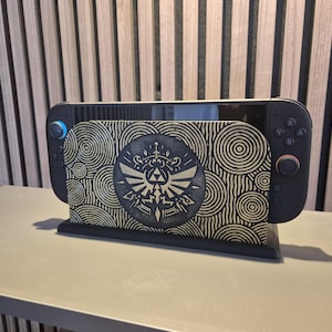Peut inclure: Support de console de jeu noir et doré avec un motif tourbillonnant et un emblème central. Il maintient une console de jeu noire avec des boutons bleus et rouges. Le design est inspiré d'un jeu d'aventure fantastique populaire.