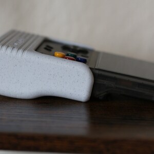 May include: A white handheld gaming console with a textured grip and a black screen. The console has a row of colorful buttons on the top.