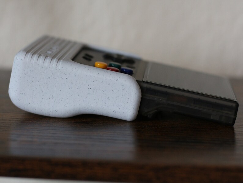 May include: A white handheld gaming console with a textured grip and a black screen. The console has a row of colorful buttons on the top.